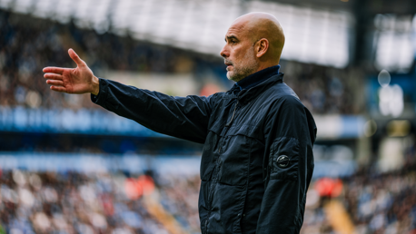 A bald man wearing a black jacket is gesturing with his hand outstretched on a football field, with stands full of spectators in the background.