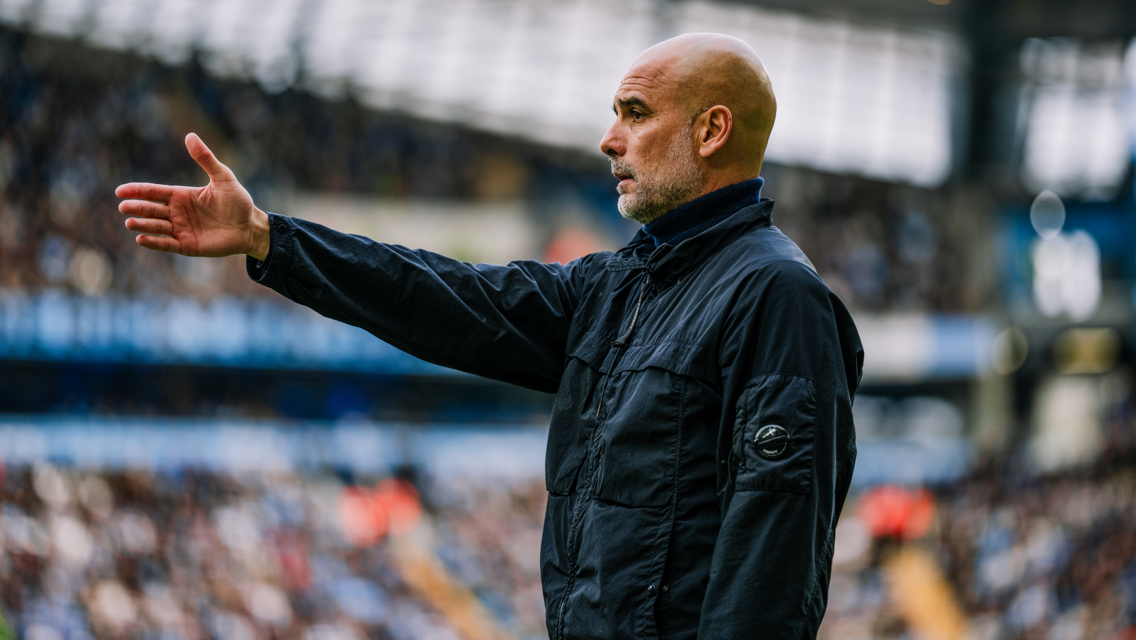 A bald man wearing a black jacket is gesturing with his hand outstretched on a football field, with stands full of spectators in the background.