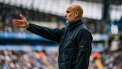 A bald man wearing a black jacket is gesturing with his hand outstretched on a football field, with stands full of spectators in the background.