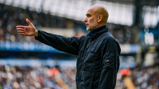 A bald man wearing a black jacket is gesturing with his hand outstretched on a football field, with stands full of spectators in the background.