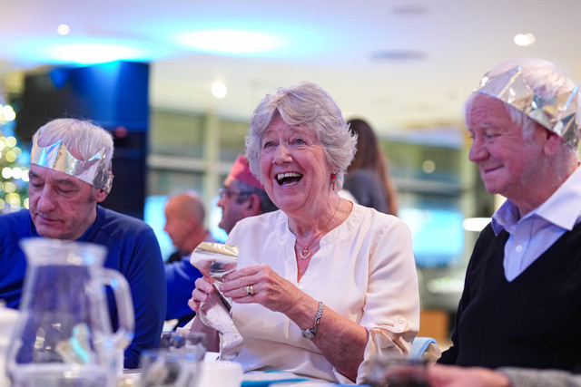 Three elderly people sitting around a table at an indoor event, with two wearing shiny hats. Their faces are obscured for privacy.