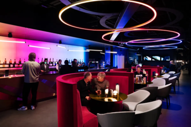 A modern lounge bar with neon lights, circular ceiling fixtures, and red seating booths. People are sitting and chatting with drinks at tables.