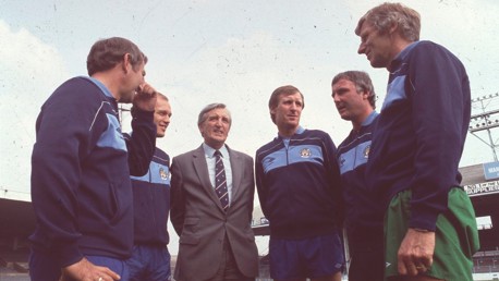 Group of men in Manchester City tracksuits standing together at a stadium, possibly discussing strategy.