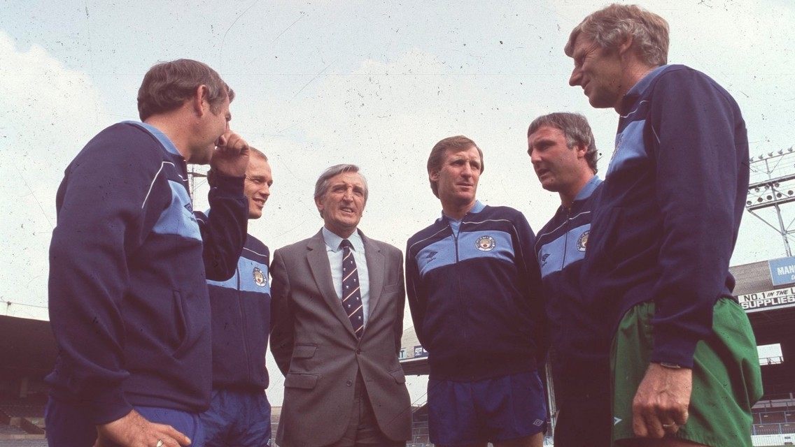 Group of men in Manchester City tracksuits standing together at a stadium, possibly discussing strategy.