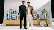 Two men standing in front of a display of various trophies, including Premier League, FA Cup, and Community Shield.