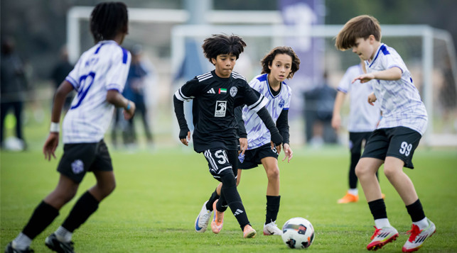 Children playing soccer on a field, with one player in a black jersey dribbling the ball and others in white jerseys defending.