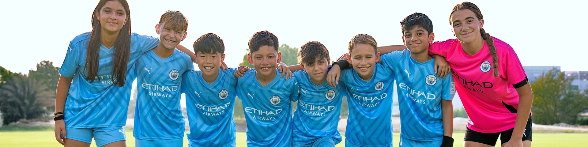 A group of young soccer players in blue Manchester City uniforms standing together on a grassy field, one in a pink goalkeeper jersey.