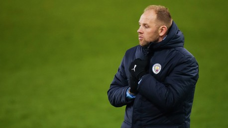 A person wearing a coat with Manchester City logo stands on a grassy field, hands together in front.