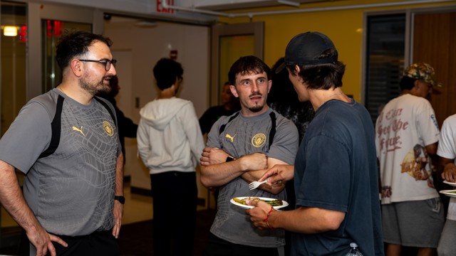 A group of people standing and talking at a social gathering, two of them wearing grey Puma shirts with a Manchester City crest.