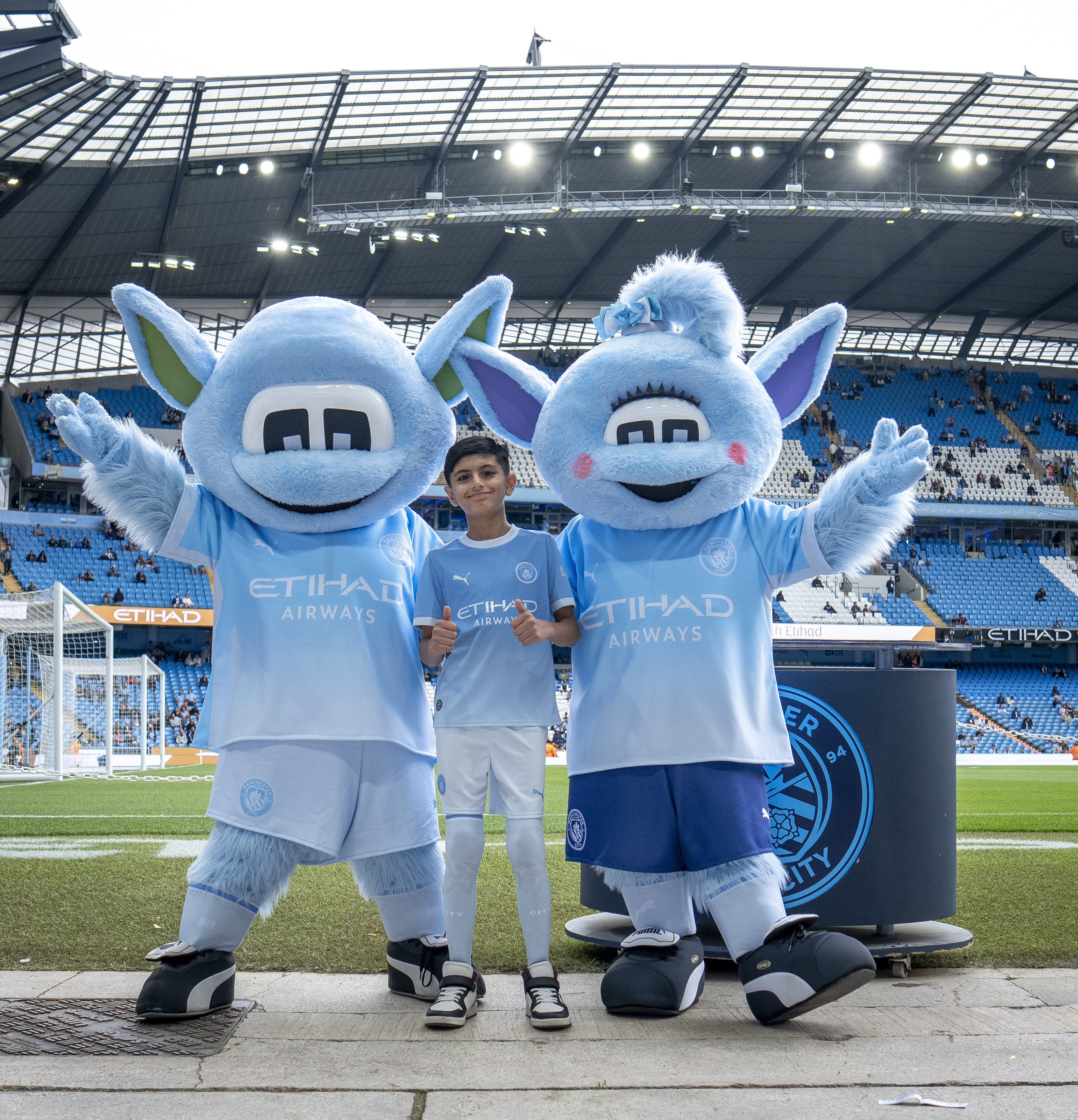A child stands between two Manchester City mascots on the field at the Etihad Stadium. The mascots are wearing Manchester City kits.
