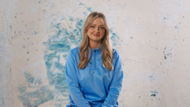 A person wearing a light blue jacket sitting against a textured blue and beige background.