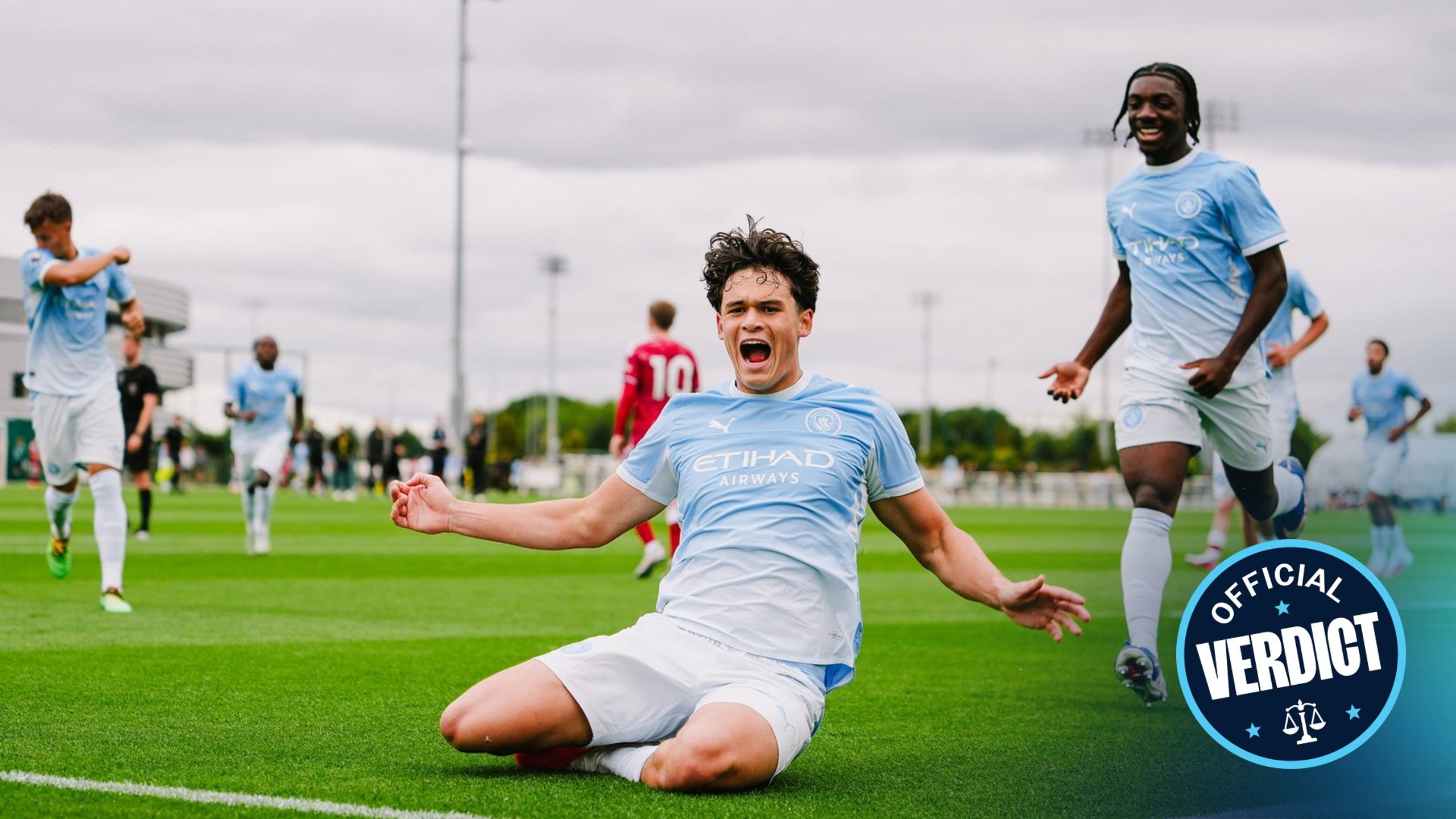 A Manchester City player celebrates a goal by sliding on the field, wearing the light blue kit. Teammates run towards him in the background. The image features a logo reading 'Official Verdict'.