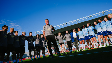 Group of people, likely a football team, standing in a circle on a field. A person in the center appears to be addressing the group. The sky is clear and blue.