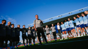 Group of people, likely a football team, standing in a circle on a field. A person in the center appears to be addressing the group. The sky is clear and blue.