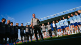 Group of people, likely a football team, standing in a circle on a field. A person in the center appears to be addressing the group. The sky is clear and blue.
