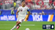 Soccer player in white and maroon jersey with number 16 dribbling on the field, wearing a blue captain's armband during a match. Stadium crowd in the background. Manchester City and World Cup logos visible.