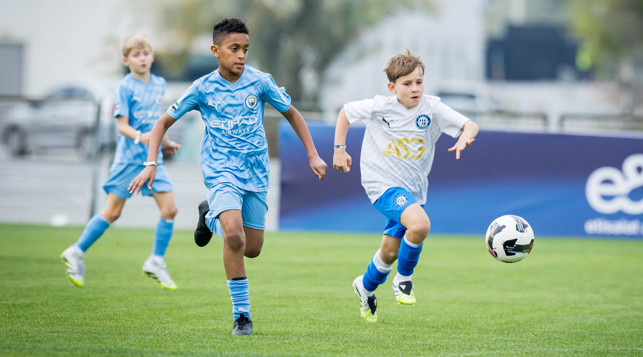 Young soccer players in blue and white jerseys engaged in a match on a grass field. The player in the blue jersey is chasing the ball along with another player in a white jersey.