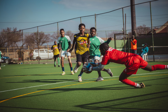 A goalkeeper in red dives to catch a soccer ball during a youth match on a green field with several players involved in the action.
