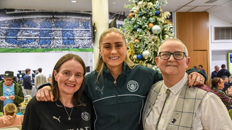 Three people standing near a decorated Christmas tree at a Manchester City gathering.