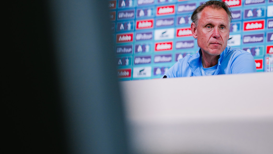 A person in a blue shirt at a press conference with a blurred face and a background of brand logos.