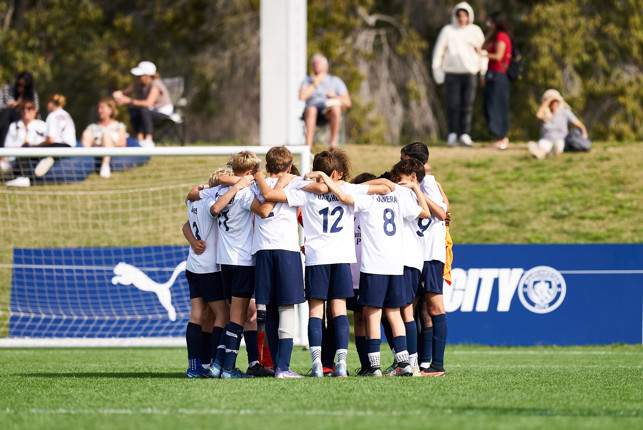 A youth soccer team in white shirts and blue shorts huddles together on the field with spectators watching in the background.