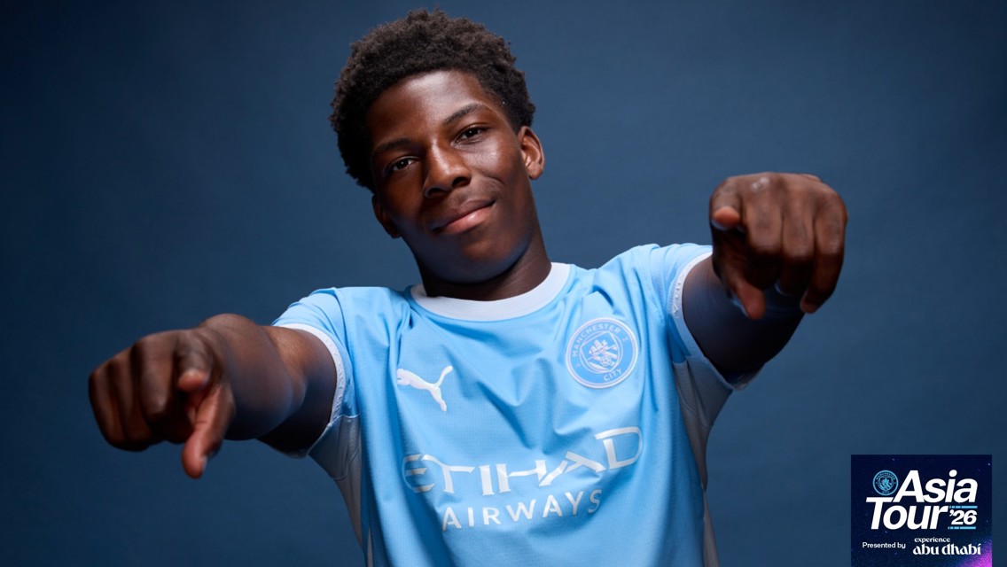 A young man wearing a Manchester City football jersey poses with both hands pointing forward. The shirt has 'Etihad Airways' and 'Puma' logos. In the corner, text reads 'Asia Tour '26' and 'experience abu dhabi'.