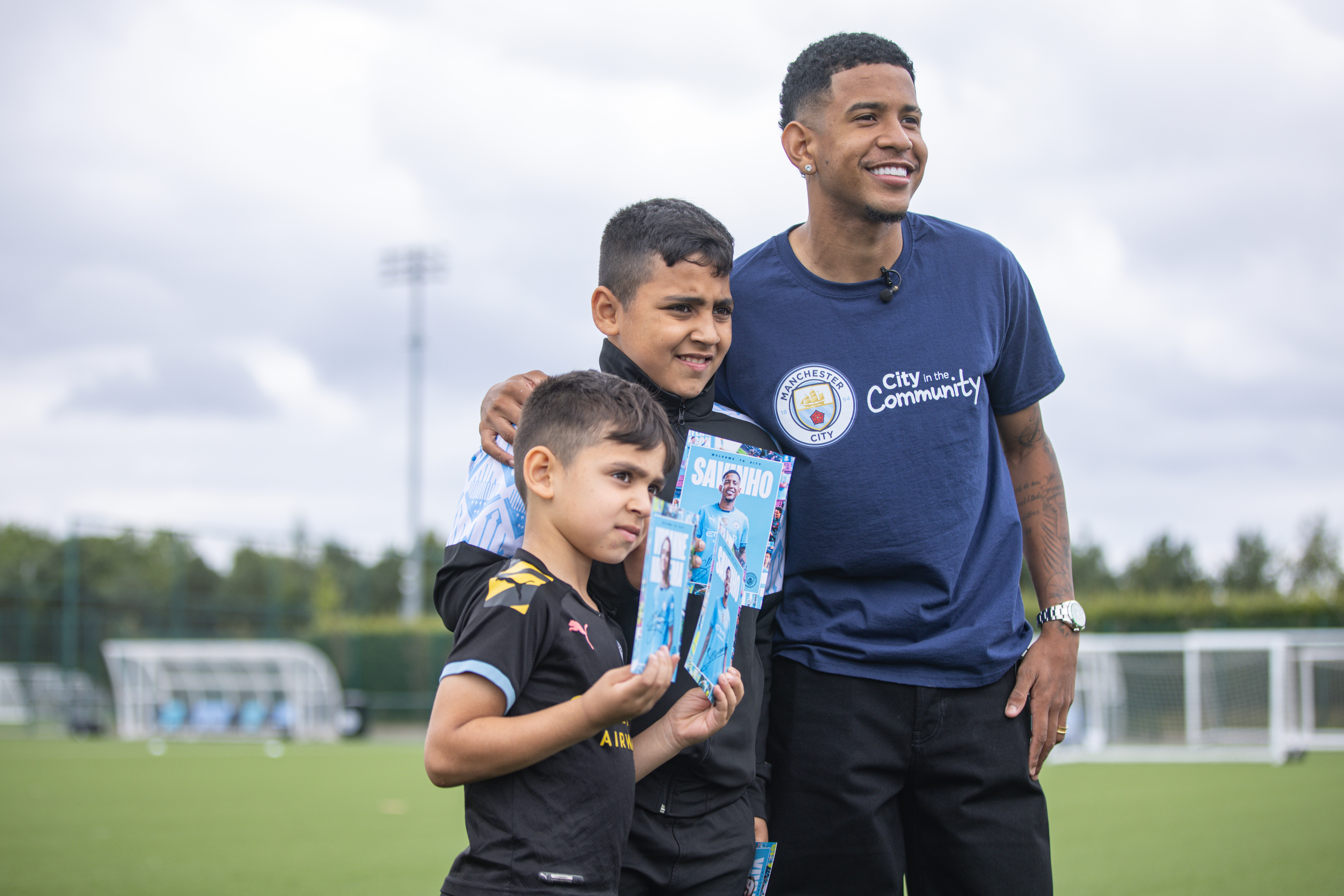 A community event photo showing a person in Manchester City Community t-shirt alongside children, with a blurred face, holding 'Sancho' comic books on a football field.