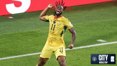 A soccer player in a yellow jersey with the number 11 is celebrating during a match. The jersey features a star and a soccer ball design, indicating a World Cup event. The background shows a green soccer field and a digital display.