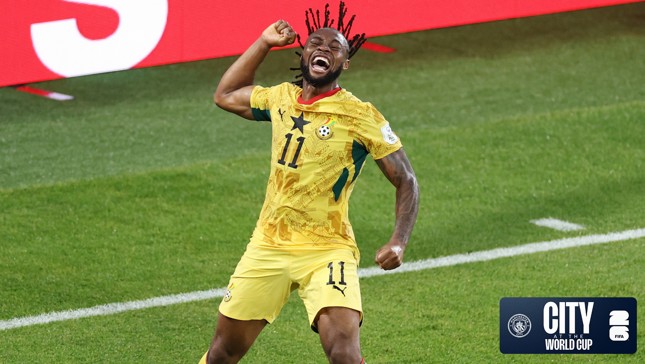 A soccer player in a yellow jersey with the number 11 is celebrating during a match. The jersey features a star and a soccer ball design, indicating a World Cup event. The background shows a green soccer field and a digital display.