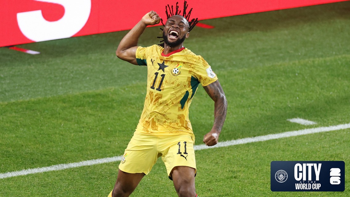 A soccer player in a yellow jersey with the number 11 is celebrating during a match. The jersey features a star and a soccer ball design, indicating a World Cup event. The background shows a green soccer field and a digital display.
