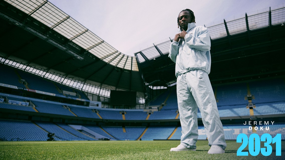 A person wearing a light blue tracksuit stands on the field inside a large stadium. The seating area is mostly empty, and the stadium has a partially open roof. Text on the image reads 'Jeremy Doku 2031'.