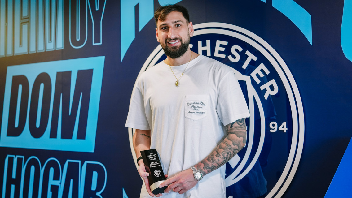 A person holding a trophy standing in front of a Manchester City logo.