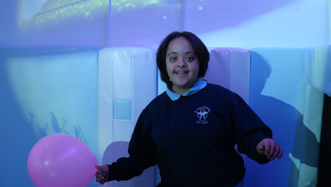 Child in a dark room holding a pink balloon with blue lighting effects on the walls.