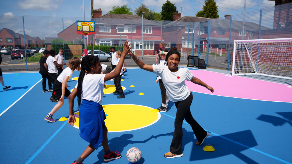 Children wearing sports attire high-five each other on a colorful outdoor sports court with cones and a soccer ball.