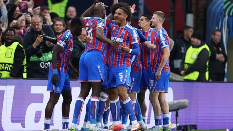 Footballers in blue and red striped uniforms celebrate a goal on the pitch. The players are surrounded by security and photographers, capturing the moment.