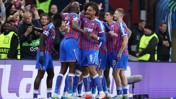 Footballers in blue and red striped uniforms celebrate a goal on the pitch. The players are surrounded by security and photographers, capturing the moment.