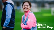 A person wearing a pink jacket with a blue vest, standing on a green field. The text 'City at the Asian Cup' is visible.