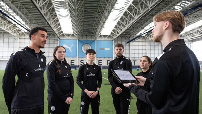 A group of individuals wearing black Manchester City training gear stands in a sports facility, with one person holding a tablet.