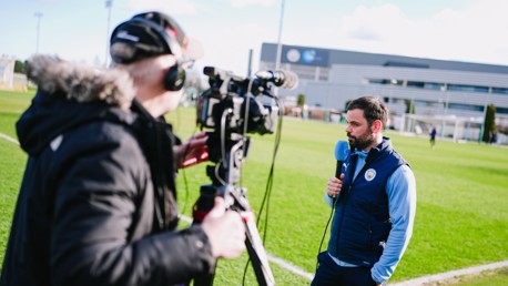 A cameraman filming a person holding a microphone wearing Manchester City attire on a football field, presumably conducting an interview.