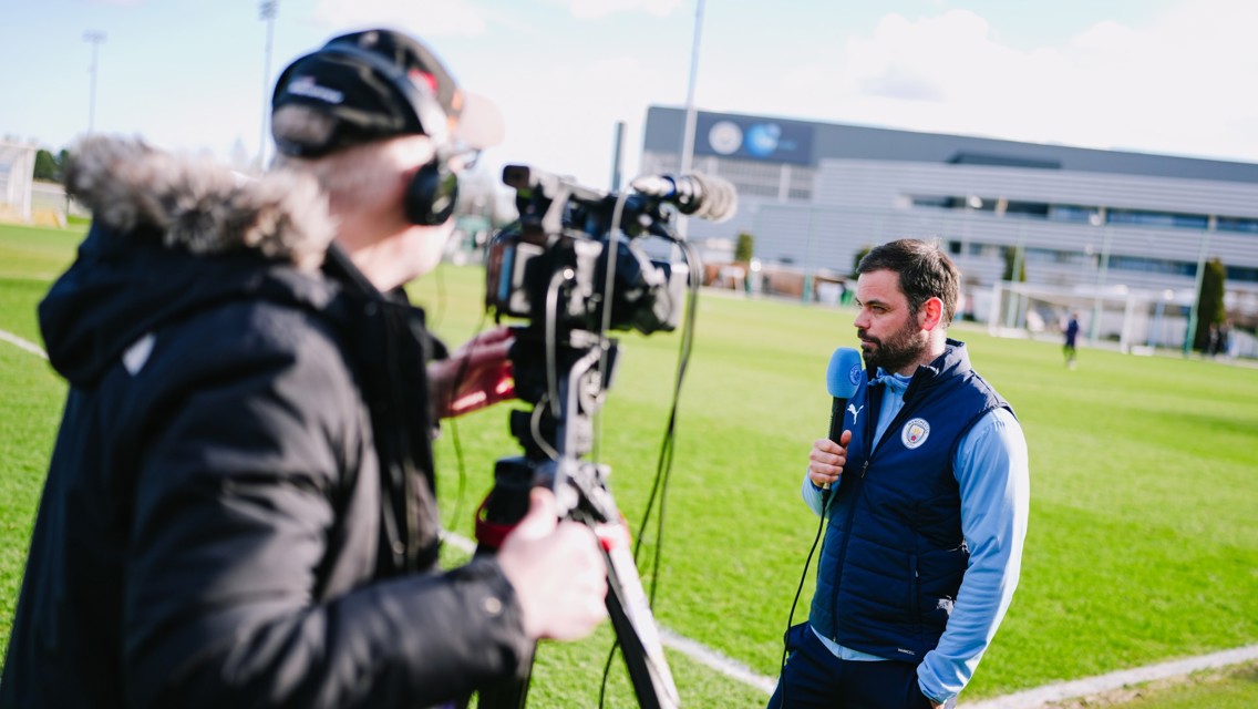 A cameraman filming a person holding a microphone wearing Manchester City attire on a football field, presumably conducting an interview.