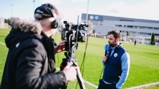 A cameraman filming a person holding a microphone wearing Manchester City attire on a football field, presumably conducting an interview.