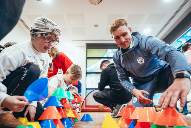 A community engagement activity with Man City personnel assisting children in stacking colorful plastic cones indoors.