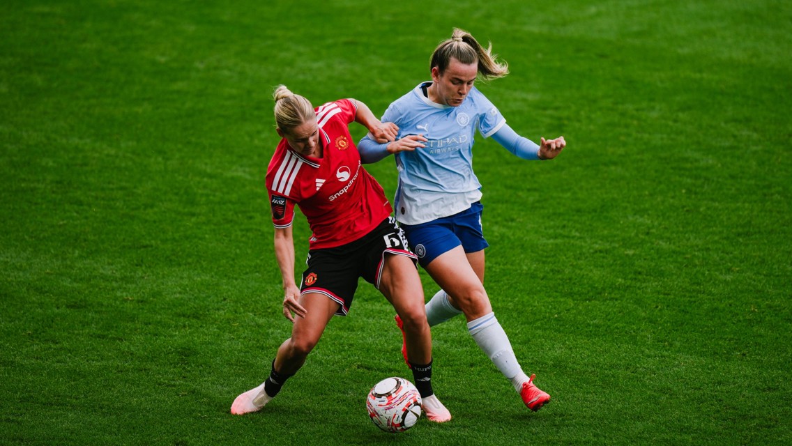 Two soccer players from opposing teams, wearing red and blue, competing for the ball during a match on a grass field.