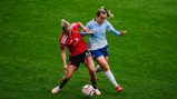 Two soccer players from opposing teams, wearing red and blue, competing for the ball during a match on a grass field.