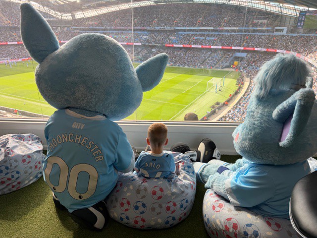 Two mascots and a child wearing Manchester City shirts sit on bean bags looking over the football field at Etihad Stadium during a game.