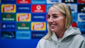 A woman in a light hoodie smiles during a press conference. Background features various brand logos.