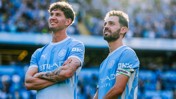 Two Manchester City players in light blue jerseys stand on the field. One player has an arm tattoo, and the other wears a captain's armband.