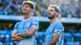 Two Manchester City players in light blue jerseys stand on the field. One player has an arm tattoo, and the other wears a captain's armband.