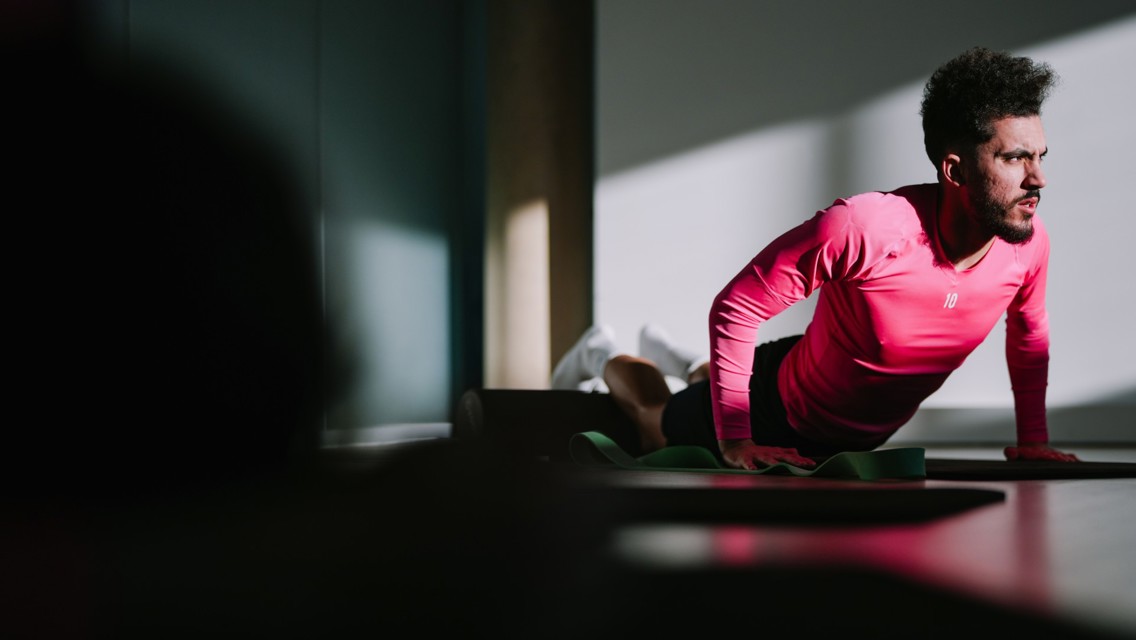 A person in a pink shirt is performing a yoga or stretching pose indoors, with sunlight streaming in.
