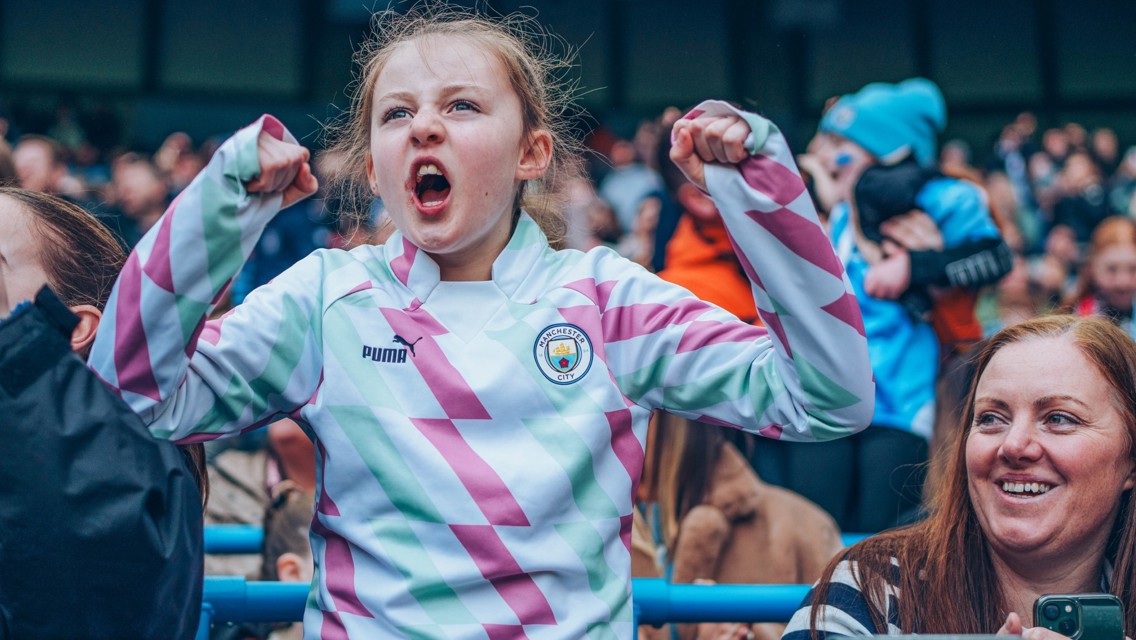 A young girl wearing a Manchester City Puma kit is seen celebrating with both arms raised in a crowd setting.